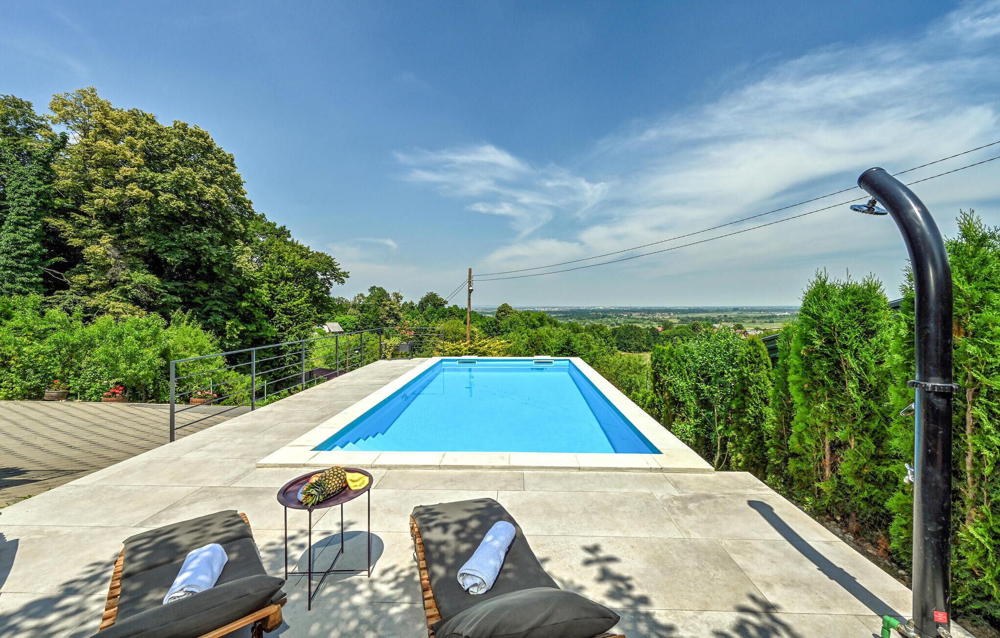 A rectangular swimming pool with a clear blue water surface, surrounded by a paved deck. There are two lounge chairs with towels, a small round table with a pineapple and a yellow cloth, and a black curved structure, possibly a water feature, to the right.