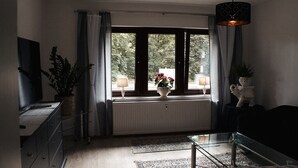 Un salón con un ventanal grande, un radiador, una mesa de centro de vidrio y un televisor sobre un mueble.