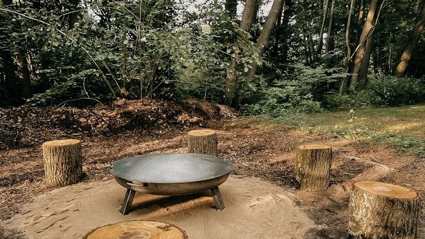 Un entorno rústico al aire libre con una olla de metal sobre una base de arena, rodeado de tocones de árboles.