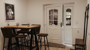 Un comedor con una mesa de madera y cuatro sillas, una puerta blanca con paneles de vidrio, y una mesita con una silla a un lado.