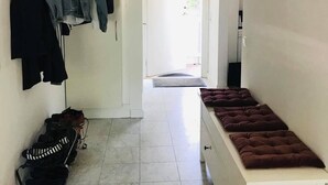 Un couloir avec un banc, un porte-manteau sur lequel sont suspendus des vêtements, une valise à roulettes et une porte donnant sur une autre pièce.