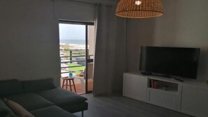 A modern living room with a large sofa, a flat-screen TV on a white cabinet, and a view of the outdoors through a sliding glass door.