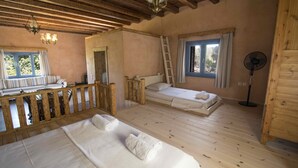 Een kamer met houten vloeren en een videbed met witte beddengoed. Er is een ladder naar een videbed en een staande ventilator bij een raam.