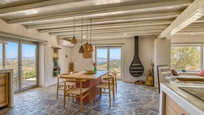 A spacious room with a stone floor, a wooden dining table with chairs, a large window with a view of the countryside, and a stone fireplace. - La Bergerie Dantiparos (Antiparos)