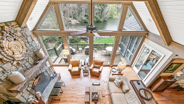 A spacious loft with a high ceiling, a stone fireplace, and a view of a pond through large windows. - Beautiful lakefront home nestled into a cove (Lake Toxaway)