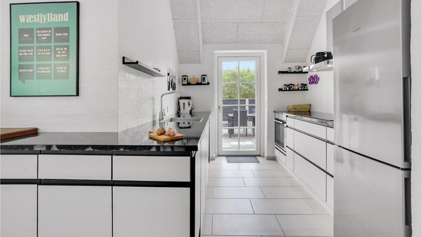 Una cocina moderna con armarios blancos, encimera negra y un refrigerador grande. Se observa un menú enmarcado en la pared y una ventana con vistas a un patio.