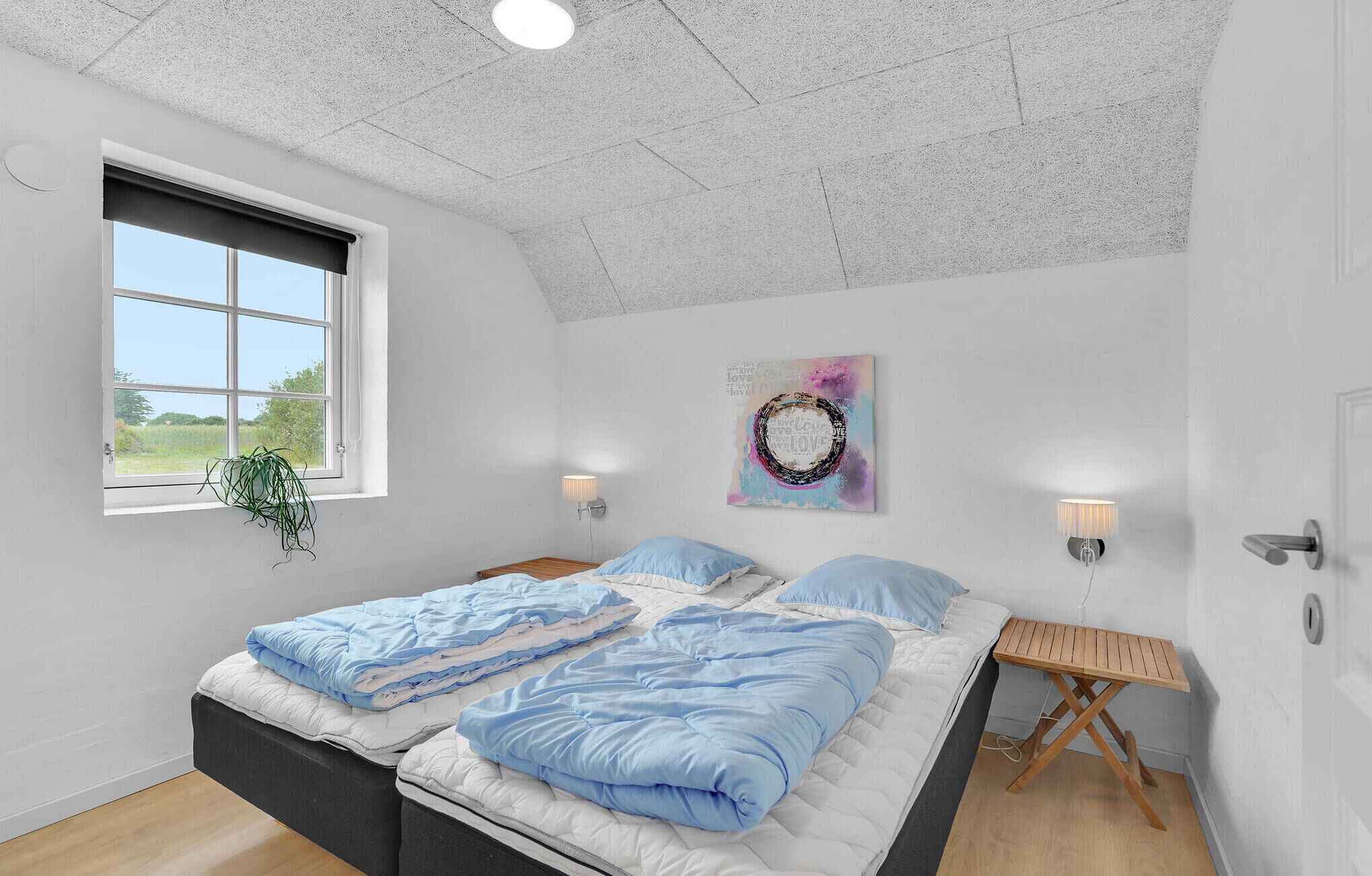 A bedroom with a large bed, a small table, a wall-mounted lamp, and a window with a view of greenery.