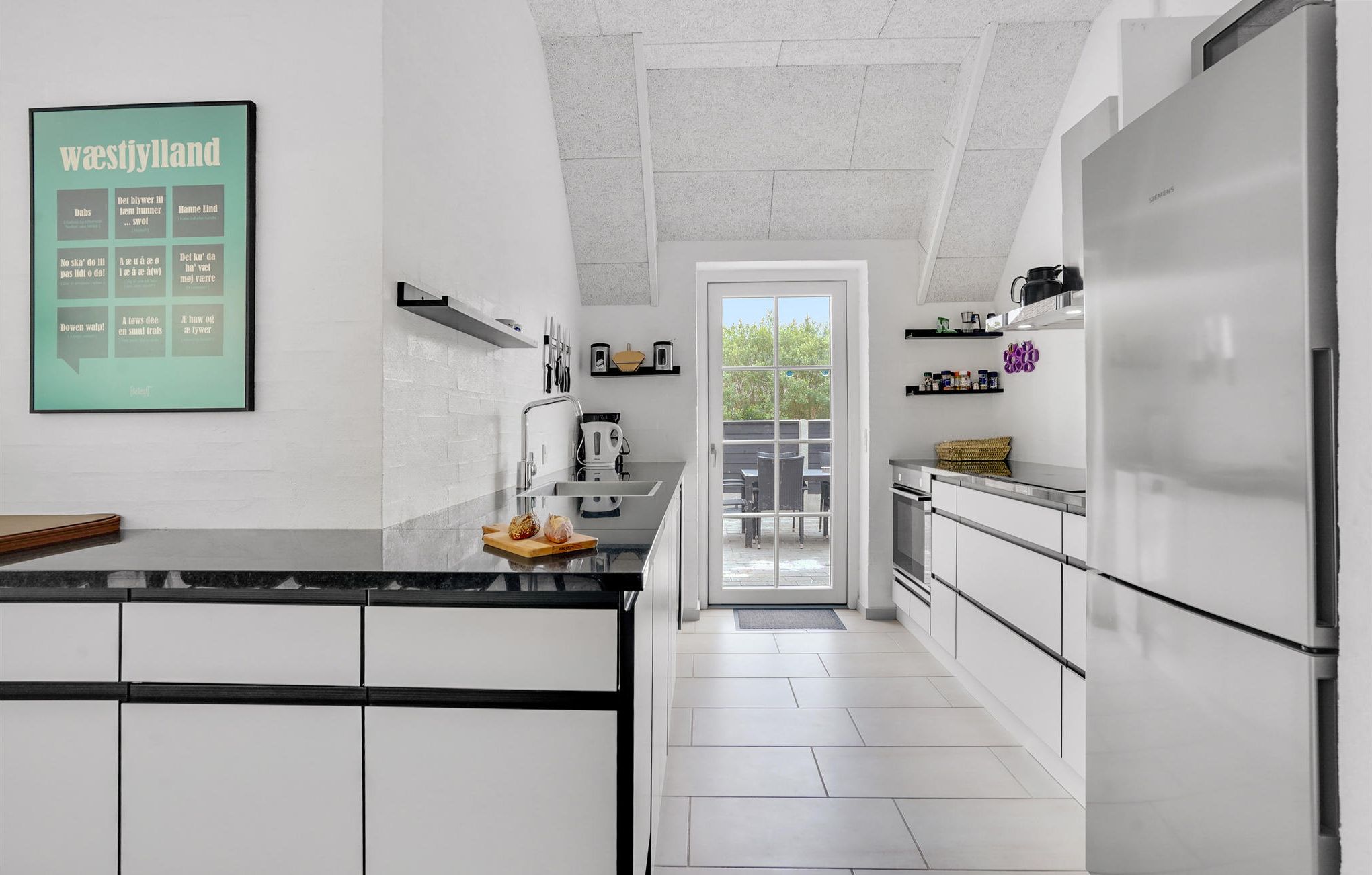 Eine moderne Küche mit weißen Schränken, einer schwarzen Arbeitsplatte und einem großen Kühlschrank. An der Wand hängt eine Speisekarte im Bilderrahmen, und ein Fenster bietet Blick auf eine Terrasse.