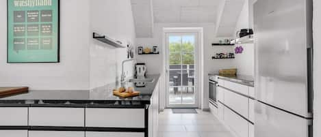 Eine moderne Küche mit weißen Schränken, einer schwarzen Arbeitsplatte und einem großen Kühlschrank. An der Wand hängt eine Speisekarte im Bilderrahmen, und ein Fenster bietet Blick auf eine Terrasse.