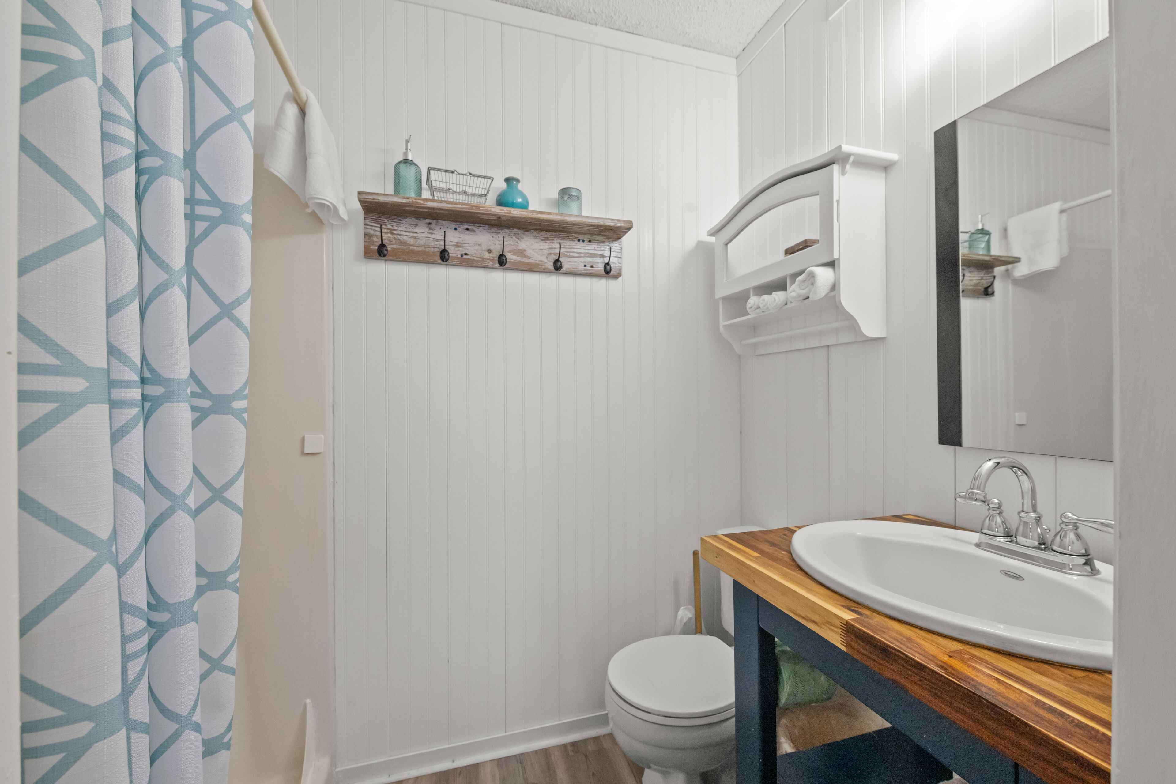 Een badkamer met een wit toilet, een houten wastafelmeubel met een wastafel en een spiegel. Daarboven hangt een handdoekhouder en er is een plank met toiletartikelen.