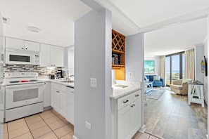 A modern kitchen with white cabinetry and a built-in microwave, leading to a living area with a sofa and a view of the outdoors.