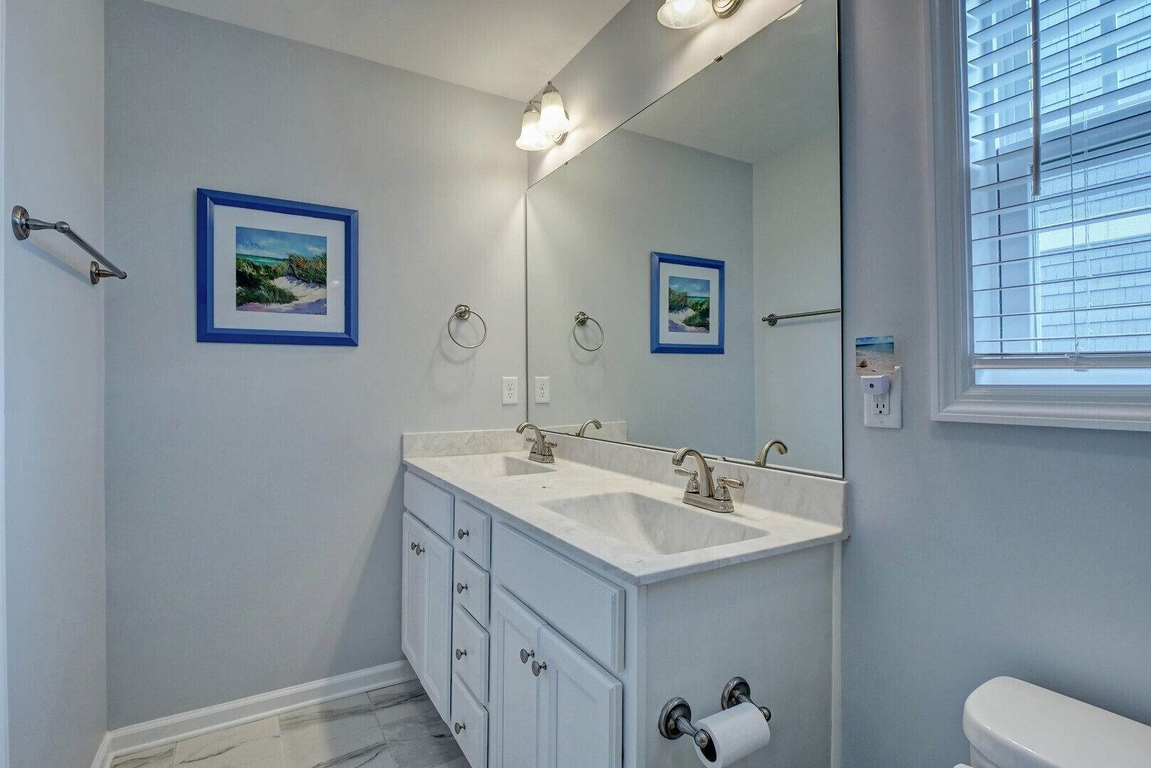 Een badkamer met twee wastafels, een grote spiegel en een toilet. Er hangen afbeeldingen in lijsten aan de muur, een handdoekhouder en een raam met zonwering.