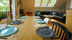 Un coin repas avec une longue table en bois, dressée pour six personnes, entourée de chaises, avec un canapé et une petite table ronde en arrière-plan.