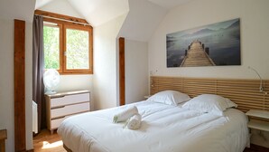 Une chambre à coucher avec une tête de lit en bois, un grand lit avec du linge de lit blanc, une table de chevet avec une lampe, une fenêtre avec des rideaux et un tableau représentant une jetée accroché au mur.