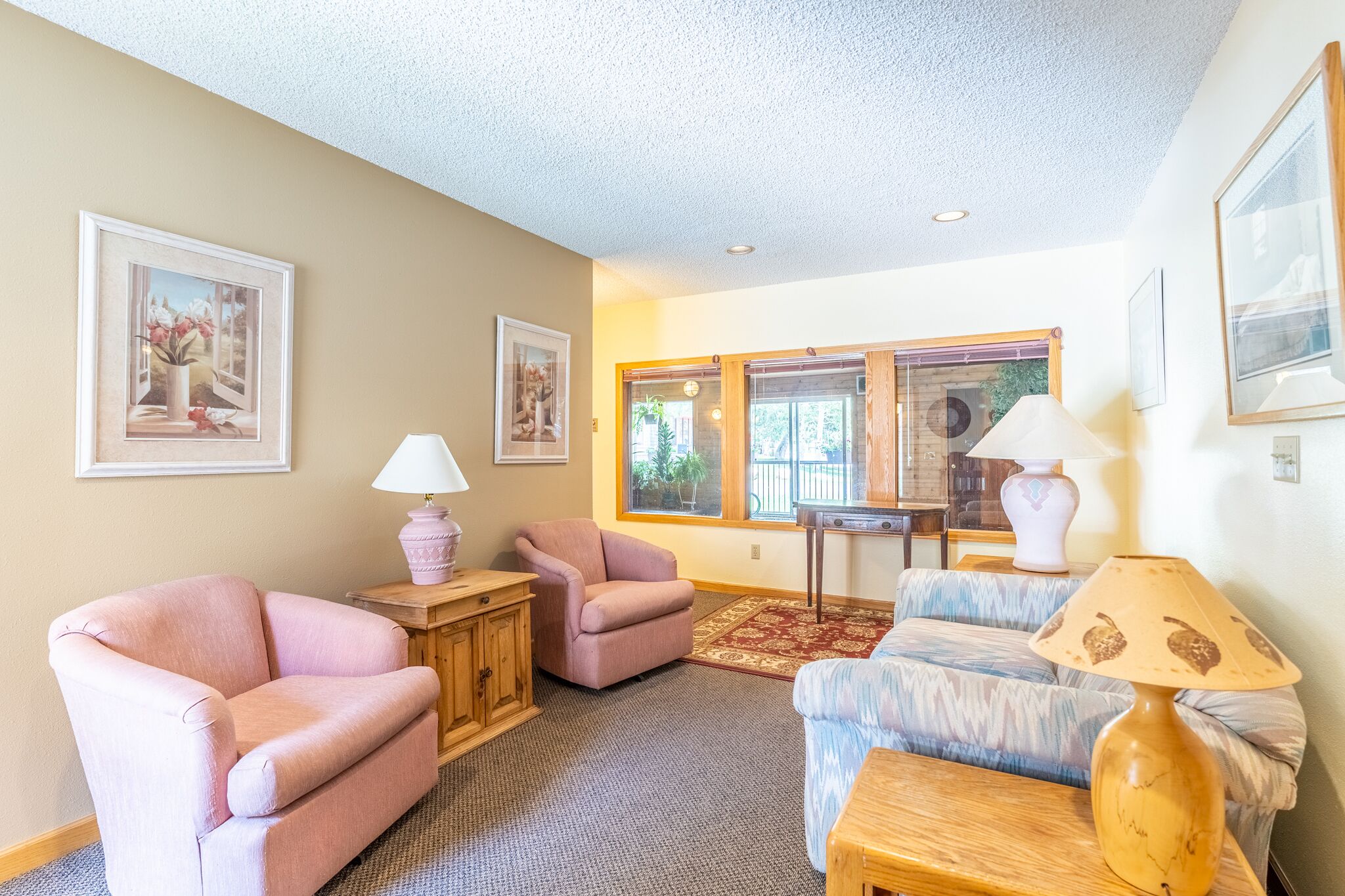 A living room with two pink armchairs, a blue sofa, a wooden coffee table, and a lamp. There are framed pictures on the walls and a window with a view of greenery.