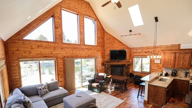 A spacious loft-style living area with a large sofa, a fireplace, and a kitchen with wooden cabinets and a stone countertop. - Edgewater Vista - Waterfront home, sleeps 6, short drive or walk to town! (Grand Lake)
