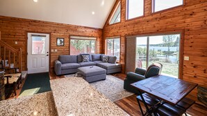 A spacious living room with a large sectional sofa, a dining table, and a view of a lake through large windows. - Edgewater Vista - Waterfront home, sleeps 6, short drive or walk to town! (Grand Lake)