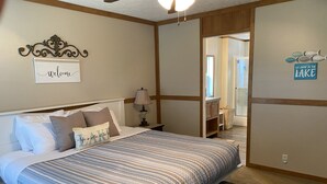 A neatly made bed with a striped bedspread, a bedside table with a lamp, and a wall sign that reads "Welcome".