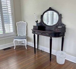 A room with a dark wooden vanity table, a white wicker chair, and a clear plastic waste basket.