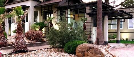A two-story house with a garden featuring palm trees, a large rock, and red flowers.