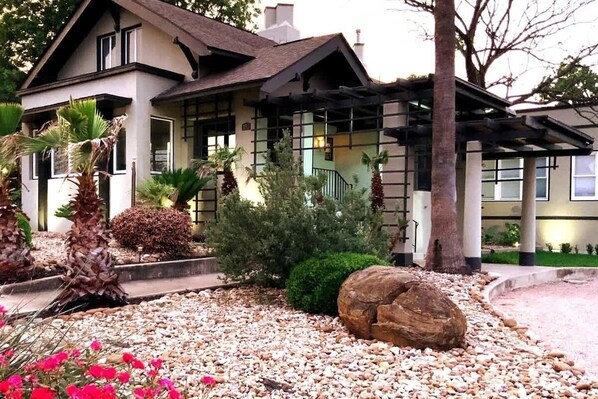 A two-story house with a garden featuring palm trees, a large rock, and red flowers.