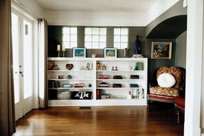 A spacious room with a wooden floor, a white bookshelf filled with books and decorative items, a patterned armchair, and a painting on the wall.