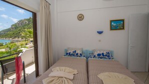 Ein Zimmer mit zwei Betten, einem Balkon mit Blick auf Strand und Berge, einem Gemälde und einer Tür.