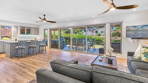 A spacious living area with a grey sofa, a coffee table, and a TV on the wall. There are ceiling fans, a kitchen with a bar area, and large sliding glass doors leading to an outdoor patio. - 100-4 Island living at its best!! (Lahaina)