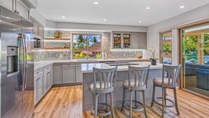 A modern kitchen with a large island, bar stools, and a view of a tropical landscape. - 100-4 Island living at its best!! (Lahaina)