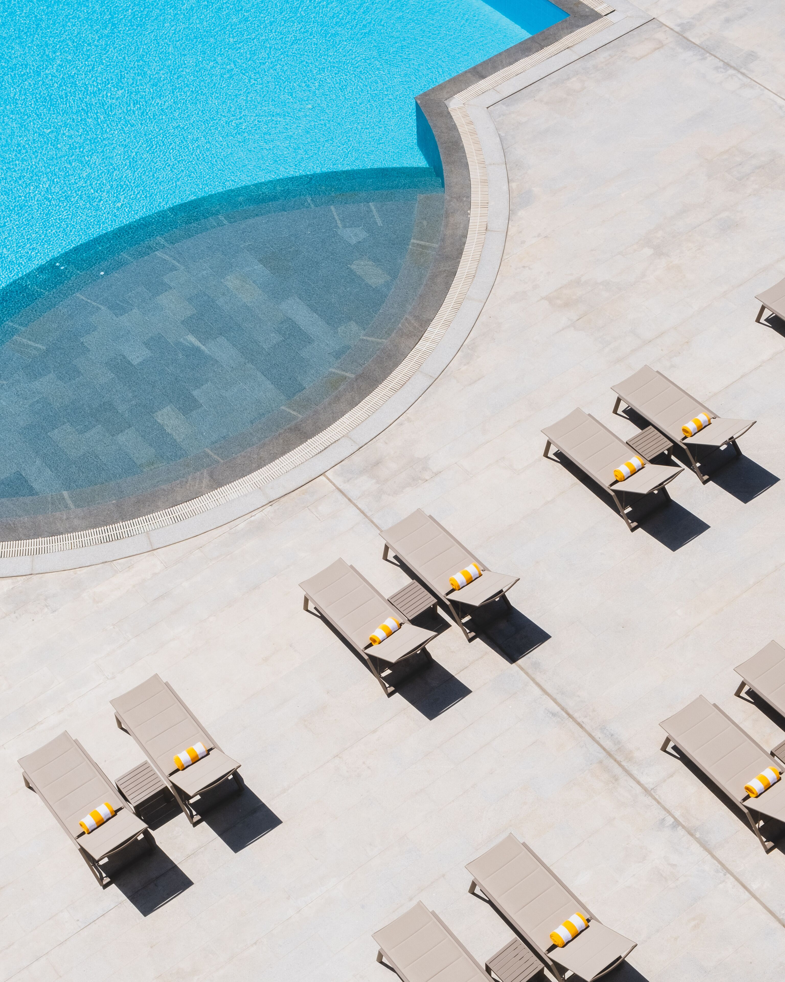 An aerial view of a pool area with a curved edge, surrounded by lounge chairs and sunbeds with yellow and white striped cushions.