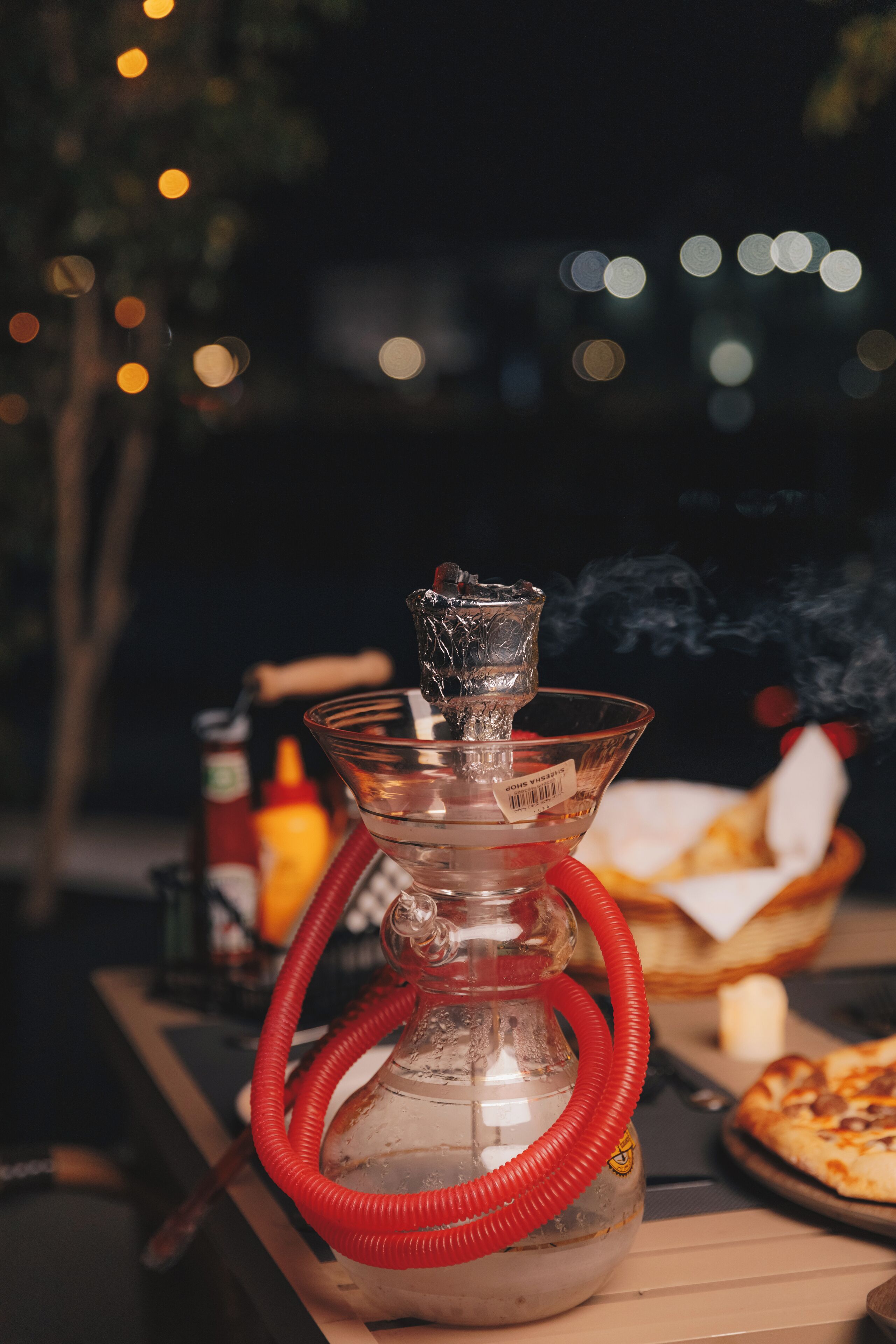 Eine Shisha mit einem roten Schlauch auf einem Tisch, im Hintergrund eine Pizza und Beilagen.