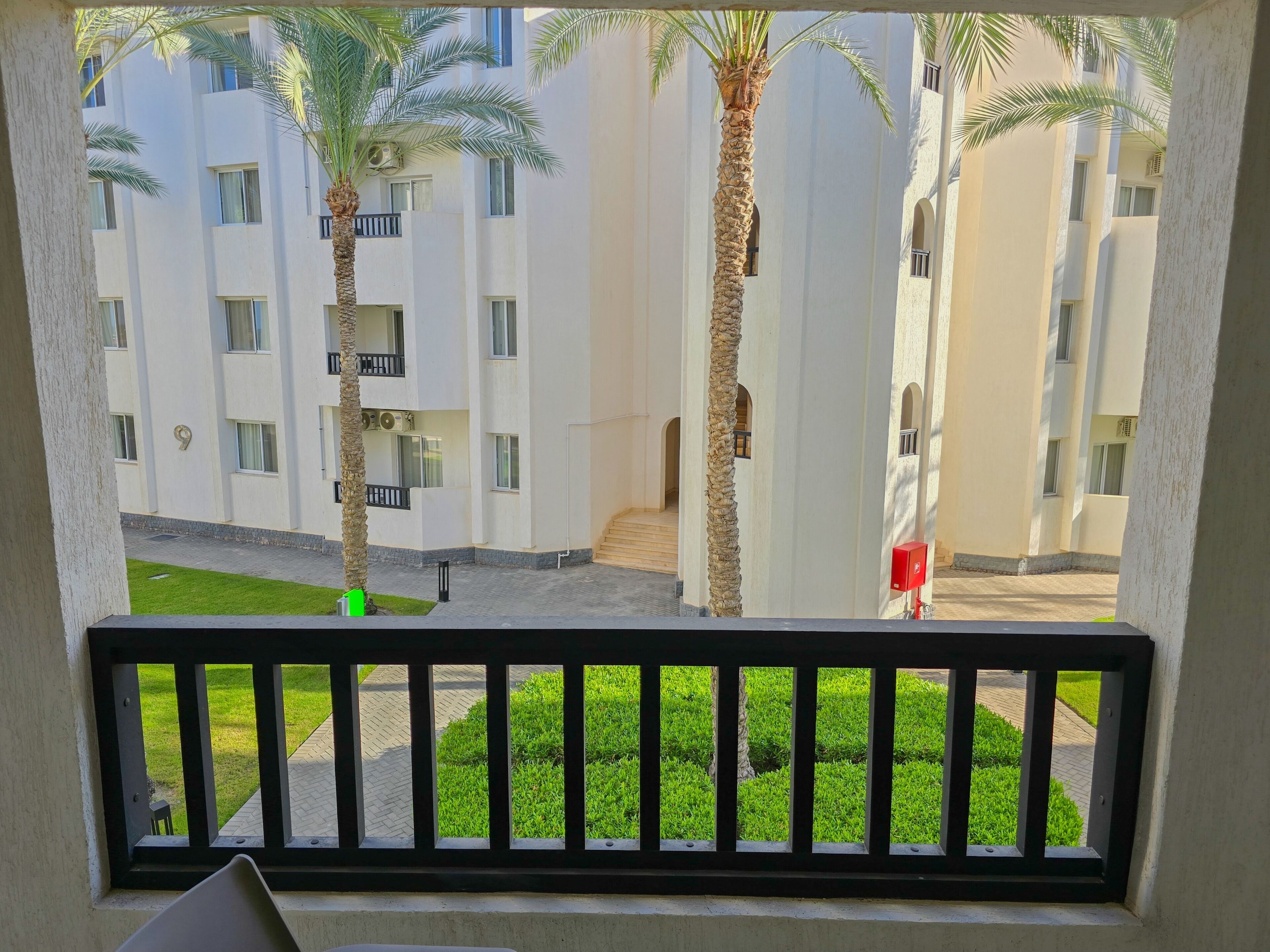A balcony with a view of a building with multiple floors, palm trees, and a well-maintained lawn.