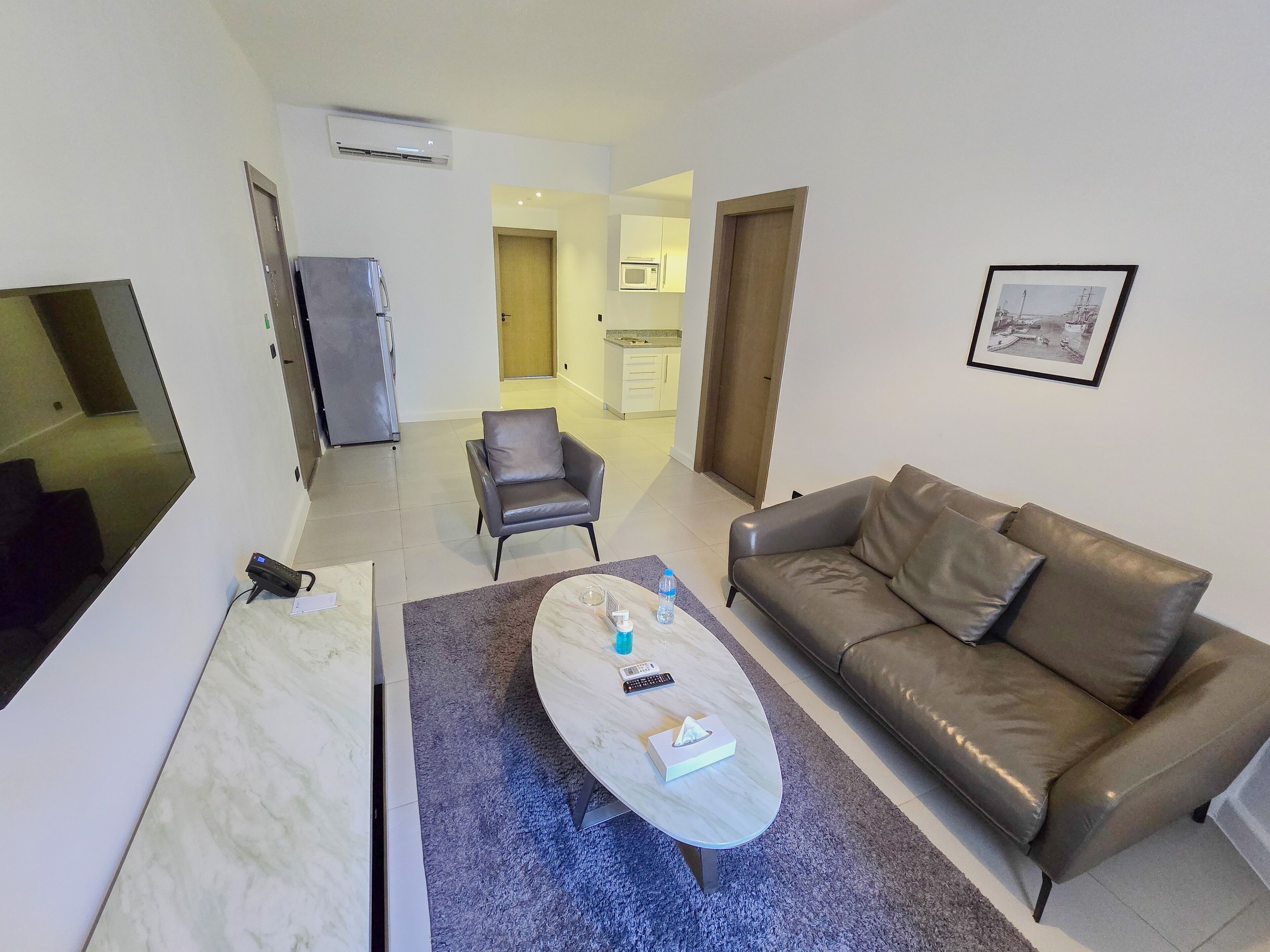 A modern living room with a sofa, a coffee table, and a chair. There is a kitchen area in the background with a microwave and cabinets.