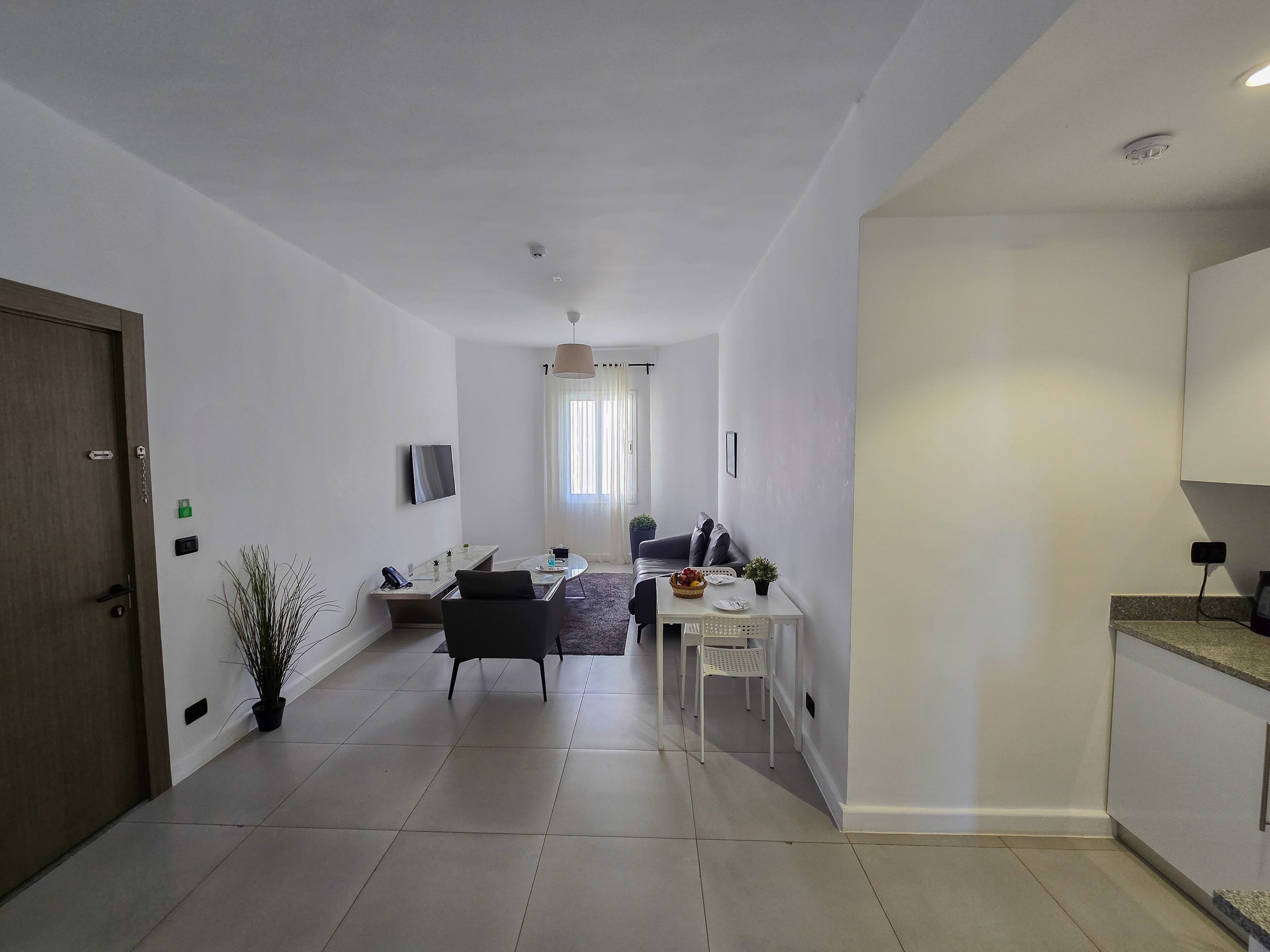Un salon moderne avec un canapé, une table basse et un téléviseur. Il y a également un coin repas avec une table et des chaises. Les murs de la pièce sont blancs et le sol est carrelé.