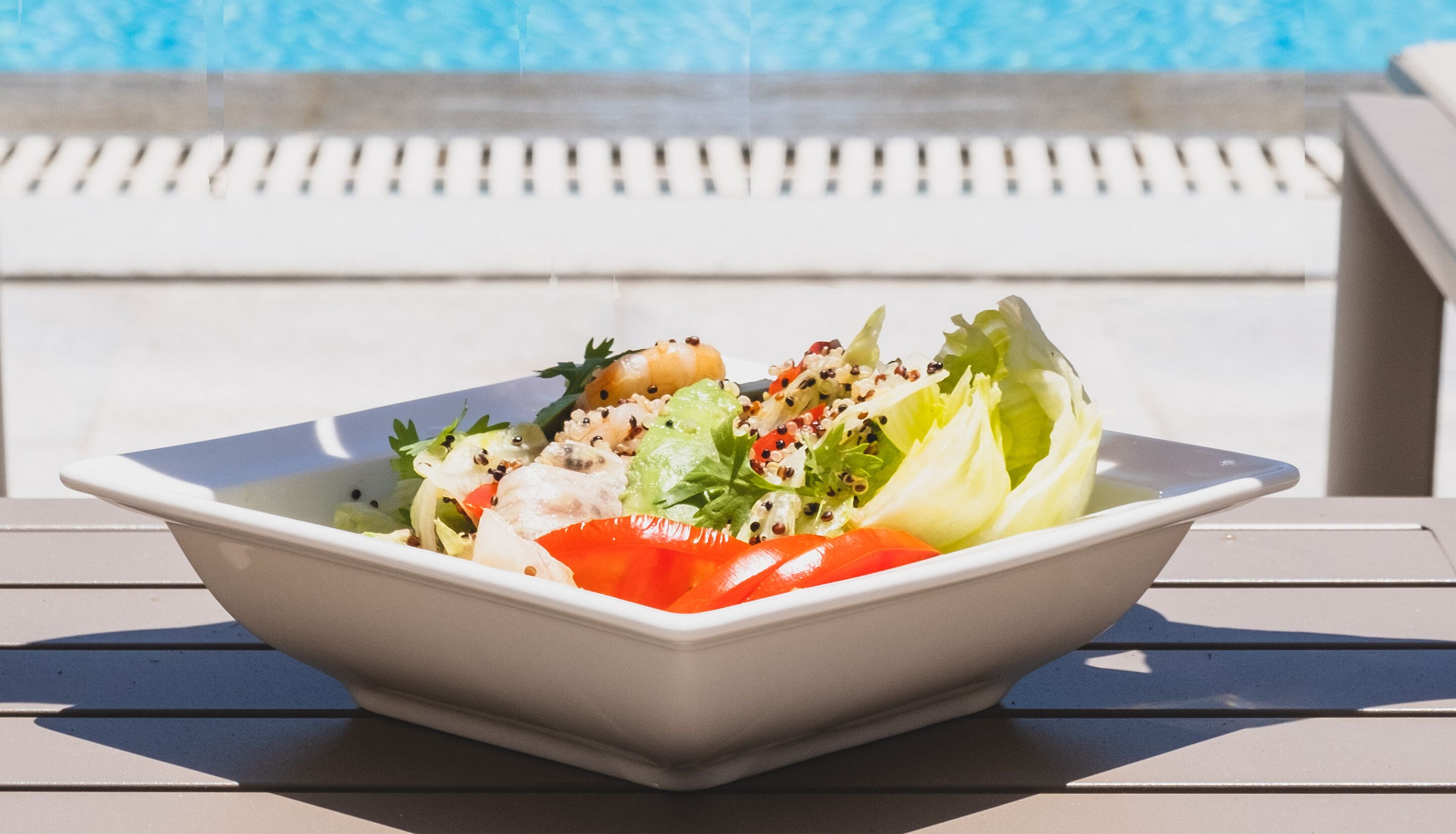 Uma tigela branca quadrada de salada com alface, tomate e outros vegetais sobre uma mesa perto de uma piscina.