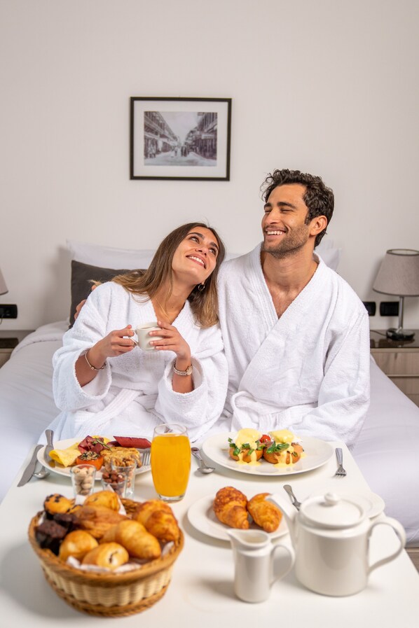 Un couple dans une chambre d’hôtel, avec un petit-déjeuner servi sur le lit, comprenant des croissants, des fruits et du jus d’orange.