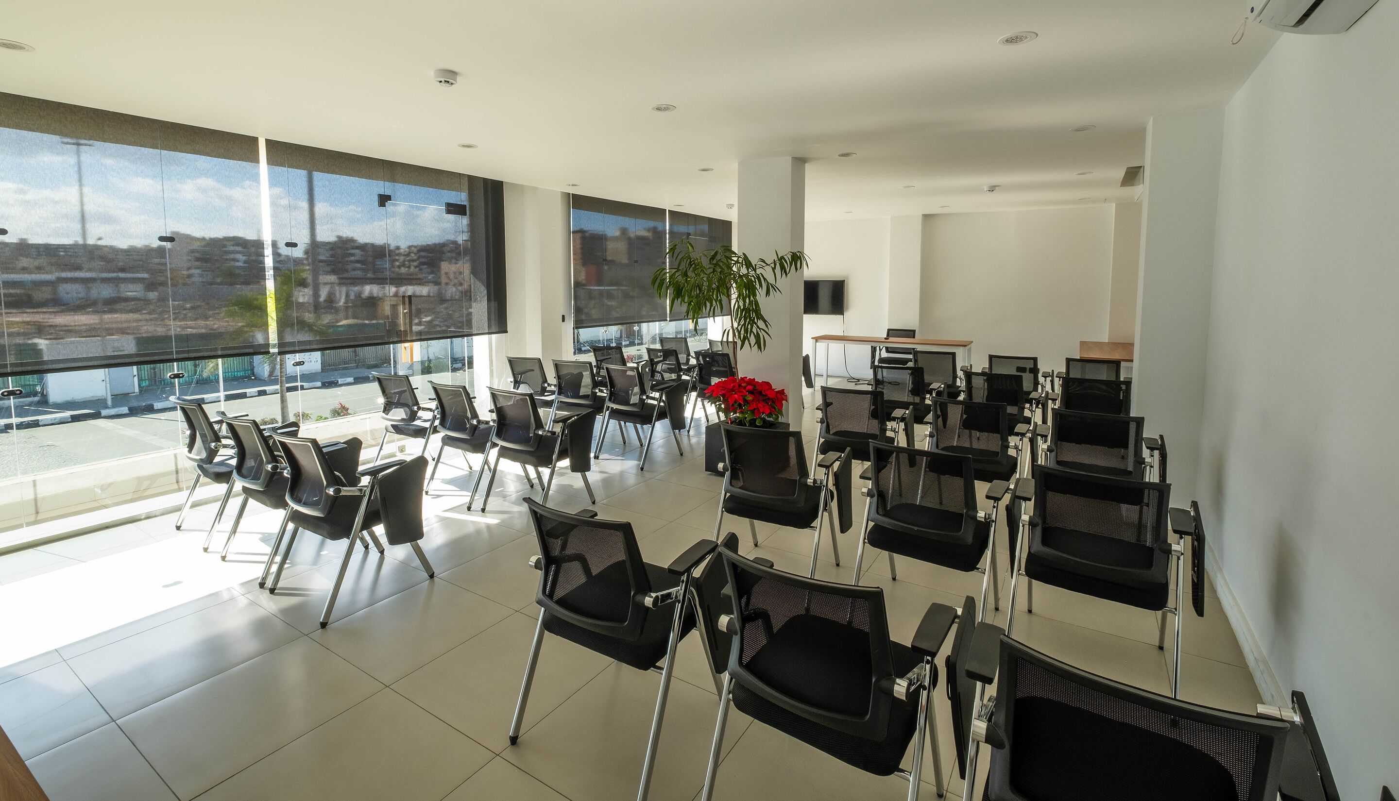 Un moderno salón de conferencias con filas de sillas negras, un amplio ventanal con vistas a la ciudad y una planta en maceta con flores rojas.