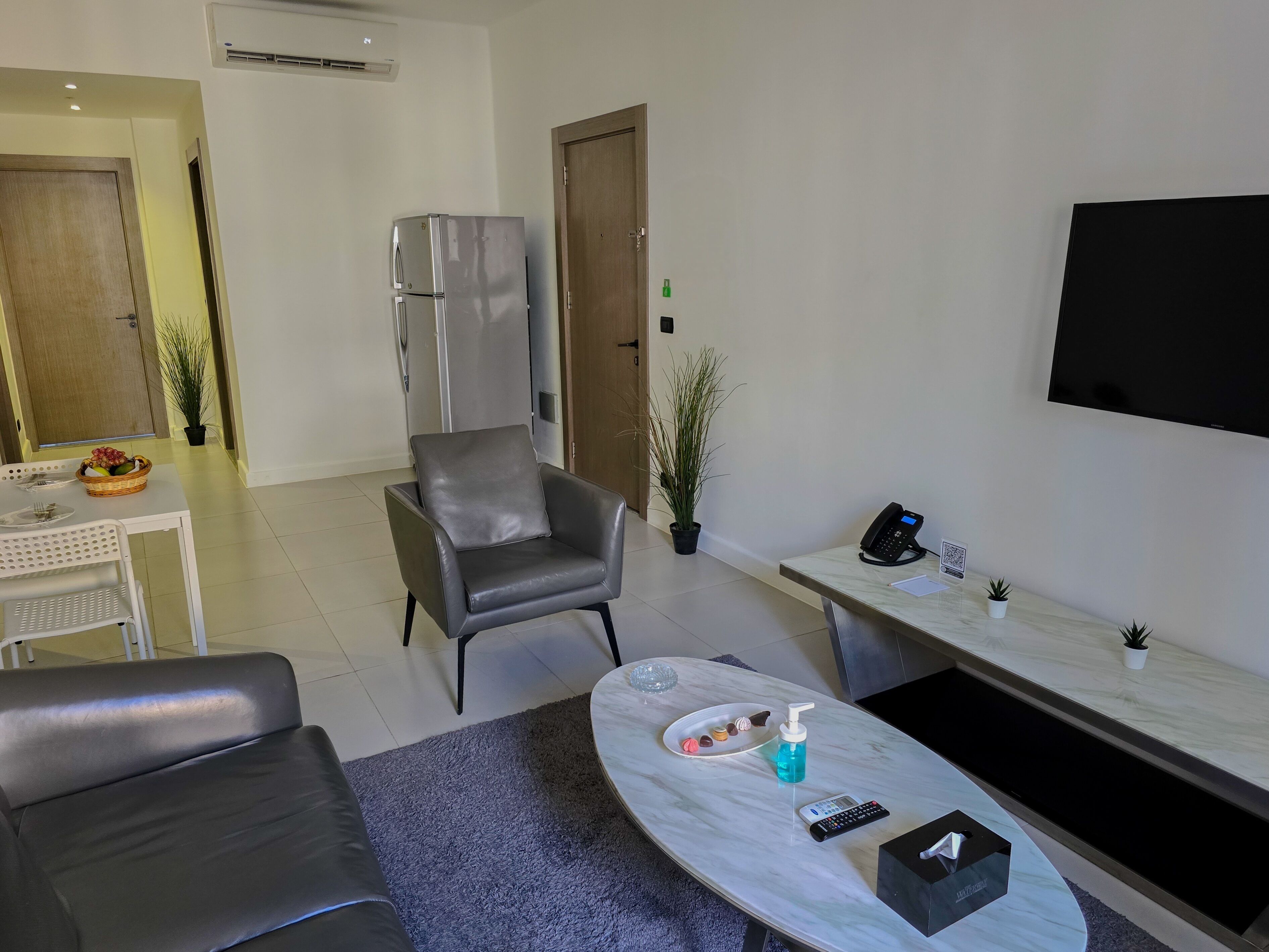 A modern living room with a grey leather sofa, a marble coffee table, a dining area with a white table and wicker basket, a grey armchair, a flat-screen TV, and a small potted plant.