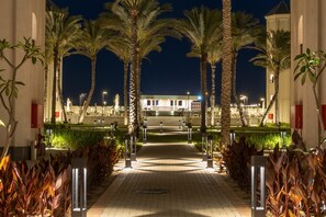 Un sendero nocturno bordeado de palmeras e iluminado por farolas, que conduce a un edificio con escalones y un césped cuidado.
