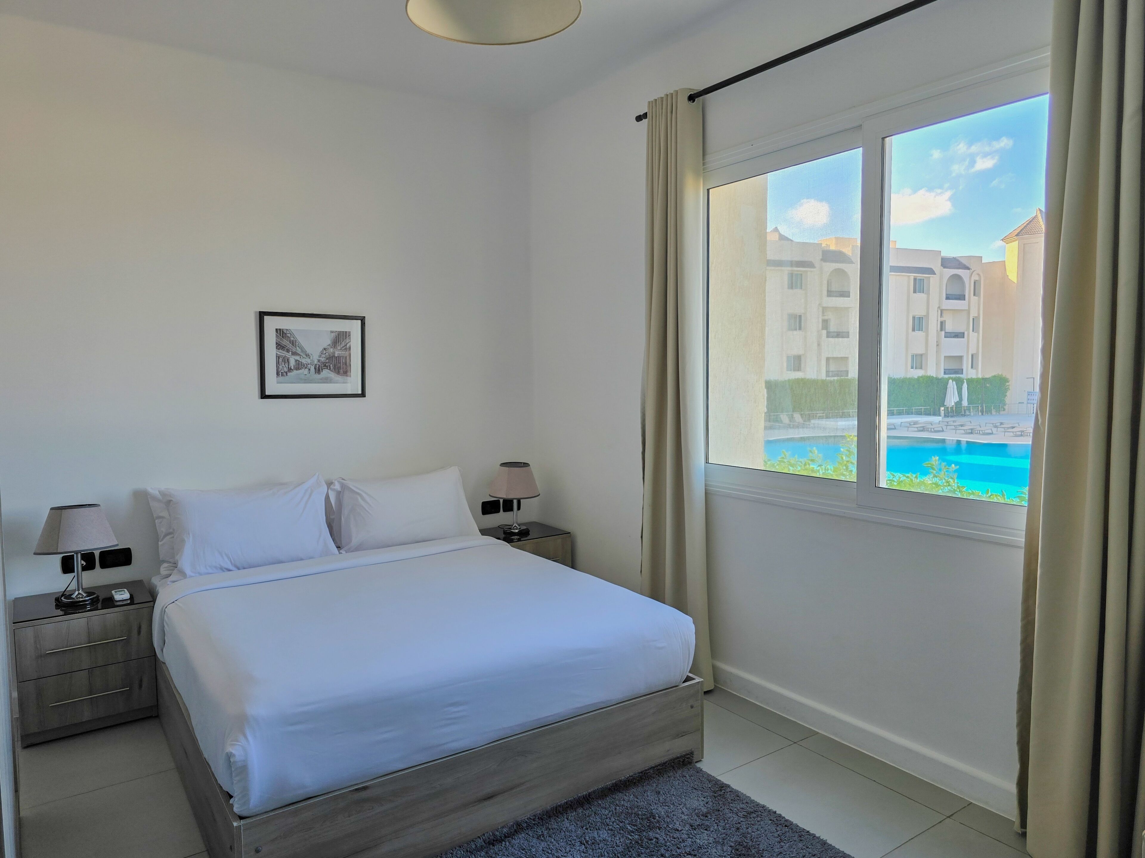 Habitación de hotel con cama, dos mesitas de noche con lámparas, un cuadro en la pared y un ventanal con vista a una piscina y edificios.