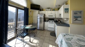 Een compacte keuken met een koelkast, magnetron en fornuis. Er is een eettafel met stoelen en een klein balkon met een stoel.