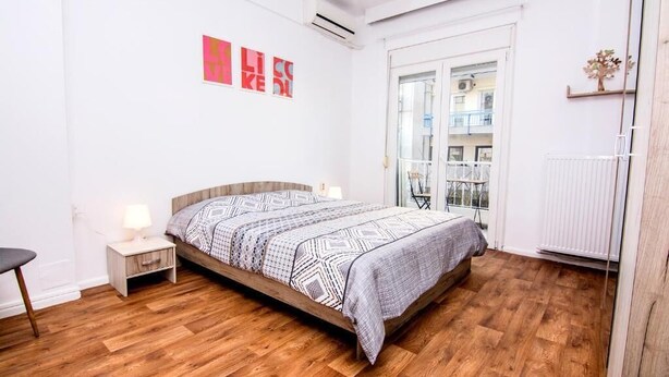 Een slaapkamer met een houten vloer, een bed met beddengoed met een patroon, een nachtkastje met een lamp, en een raam met uitzicht op het omliggende landschap.