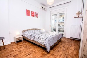 A bedroom with a wooden floor, a bed with patterned bedding, a bedside table with a lamp, and a window with a view of the outside.