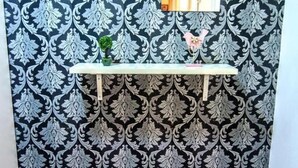 Een kamer met behang met een patroon, een witte plank met twee kleine potplanten, en twee rode krukken.
