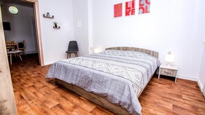 Een slaapkamer met een houten vloer, een bed met beddengoed met een patroon, een nachtkastje, een stoel en een muur met kunstwerken.