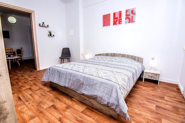 A bedroom with a wooden floor, a bed with patterned bedding, a nightstand, a chair, and a wall with artwork.