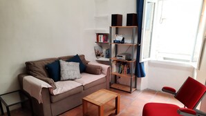Een woonkamer met een bank, een rode fauteuil, een salontafel, een boekenkast en een raam met gordijnen.
