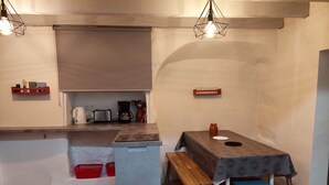 Een compacte keuken met een inbouwkoelkast, een koffiezetapparaat en een broodrooster. Er staat een houten bank en een tafel met een tafelkleed met een patroon.