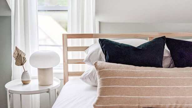 Ein ordentlich bezogenes Bett mit Holz-Kopfteil, weißer Bettwäsche und zwei dunklen Kissen.