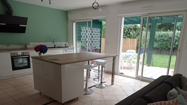 A modern kitchen with a central island, white cabinets, and a built-in oven. There is a dining area with a table and chairs, and a sliding glass door leading to a backyard with a patio and greenery. - Maison Mitoyenne Avec Ascenseur (Boucau)